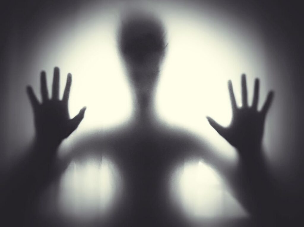 An alien or ghostlike figure is a black form behind a semi-translucent substance. A light shines from somewhere behind the creepy figure, its human-like hands pressed up against the membrane holding it back.